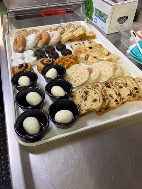 Tray with an assortment of breakfast items including hard-boiled eggs in black cups, various types of bagels, waffles, slices of bread, and pastries on a stainless steel countertop with a box of plastic wrap and some disposable face masks in the background.
