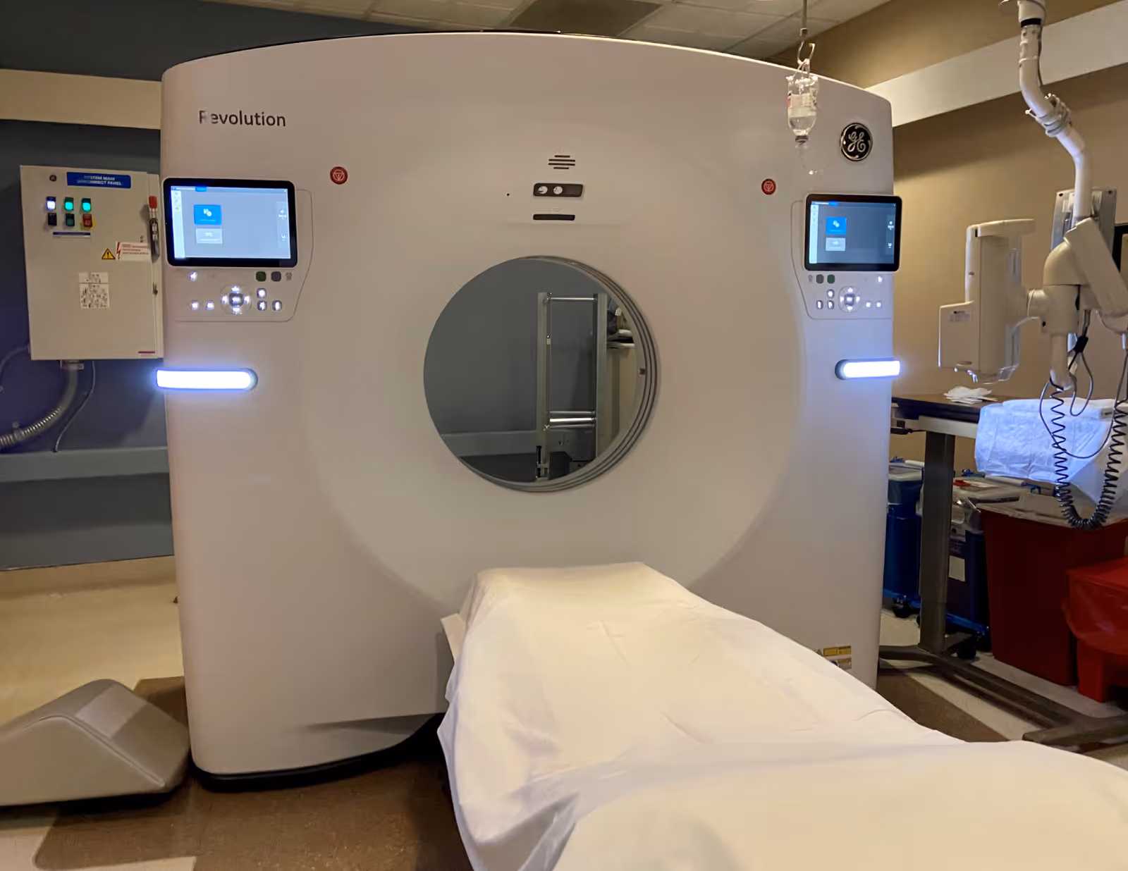 A medical imaging room featuring a large GE Revolution CT scanner with a patient table covered in a white sheet extending through the circular opening of the machine. The room has medical equipment and monitors on either side of the scanner.