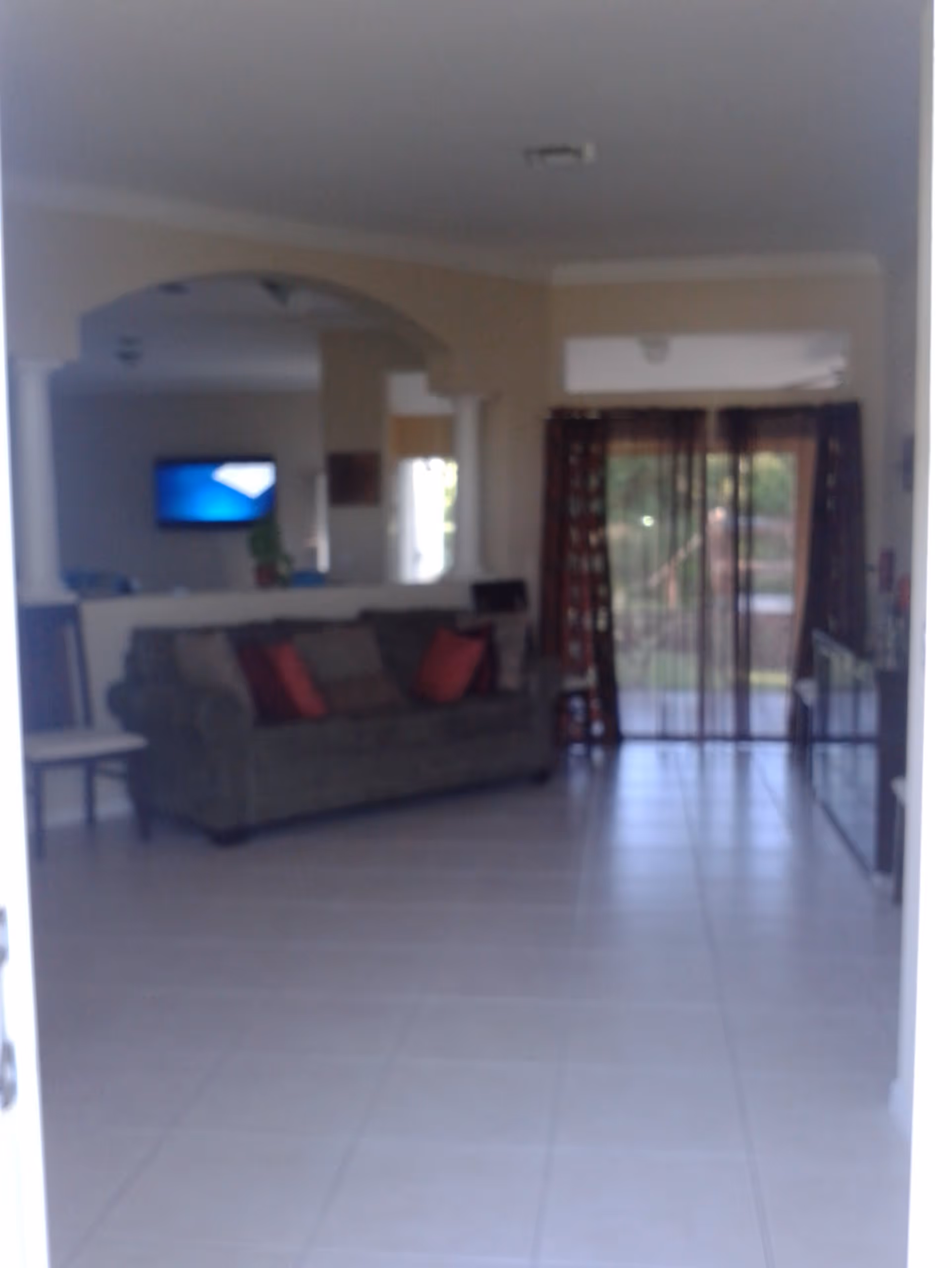 Interior view of a living room with a green couch adorned with red and brown pillows, a tiled floor, a wall-mounted TV in the background, and large glass sliding doors with sheer curtains letting in natural light.