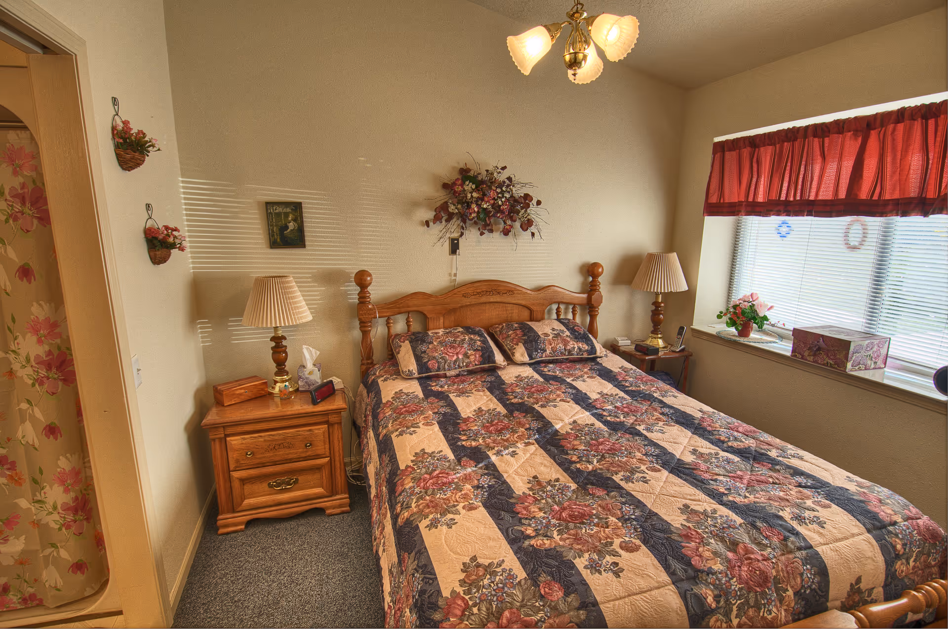 A cozy bedroom with a wooden bed frame and a floral patterned bedspread. There are two matching pillows on the bed. On each side of the bed, there are wooden nightstands with lamps. A window with red curtains lets in natural light, and there are decorative flowers on the wall above the bed and on the nightstands. A floral shower curtain is partially visible through an open doorway.