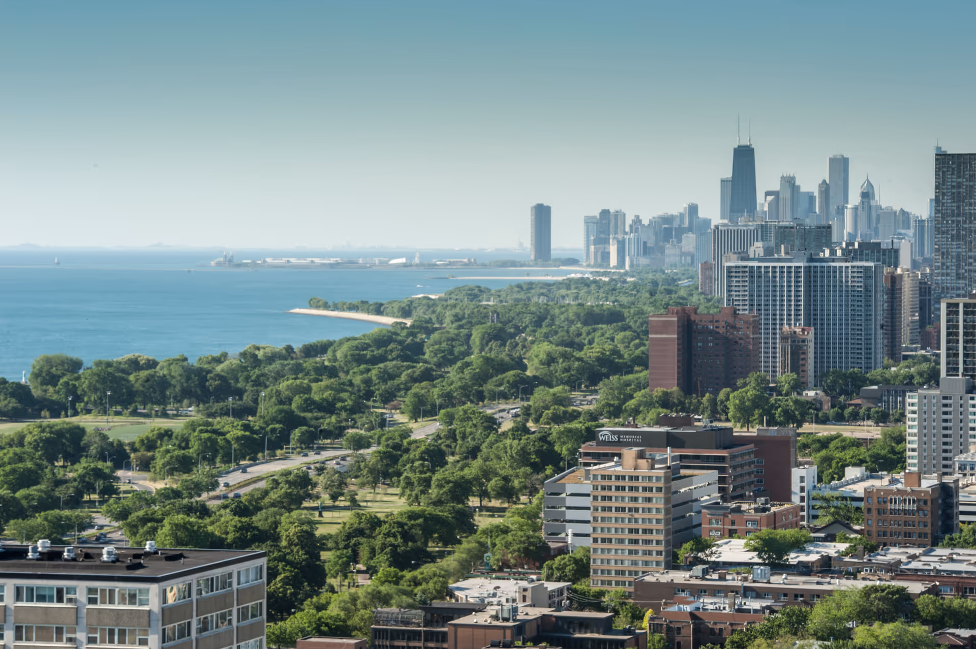 Aerial view of a city skyline with numerous tall buildings and a large green park area along the shoreline of a body of water under a clear blue sky.