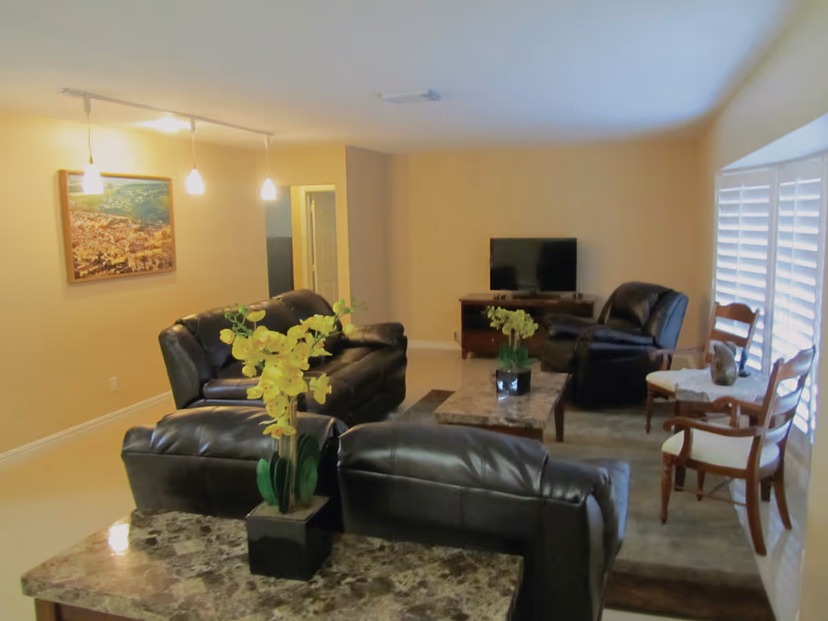 Living room with black leather sofas, a marble-top coffee table, TV on a stand, and potted flowering plants.