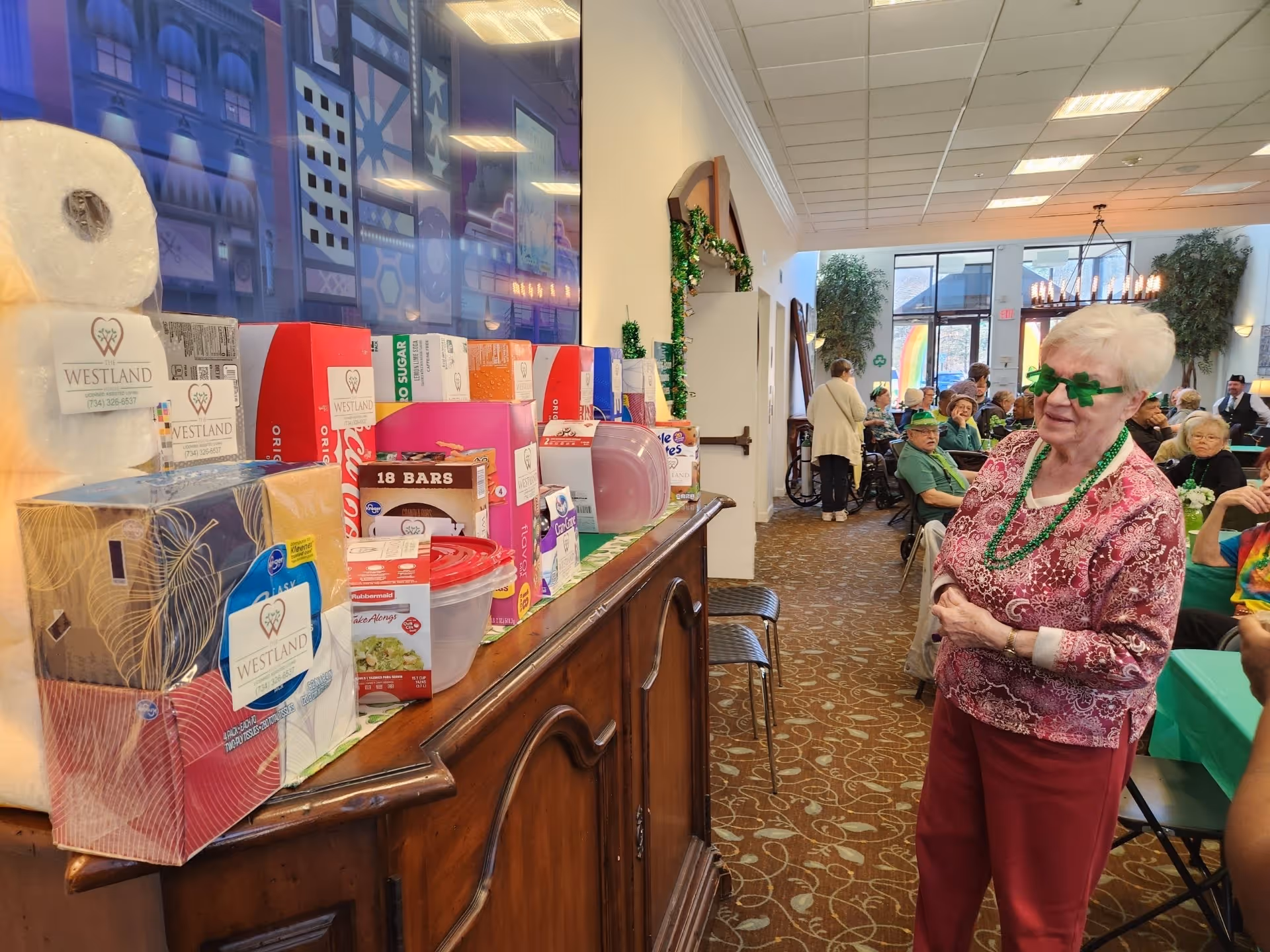 An elderly woman wearing green shamrock-shaped glasses and green bead necklaces stands in a decorated room with other seniors seated at tables in the background. On a wooden sideboard in the foreground, there are various boxed and packaged items, some labeled with 'Westland'. The room has large windows and festive decorations, suggesting a social gathering or event.