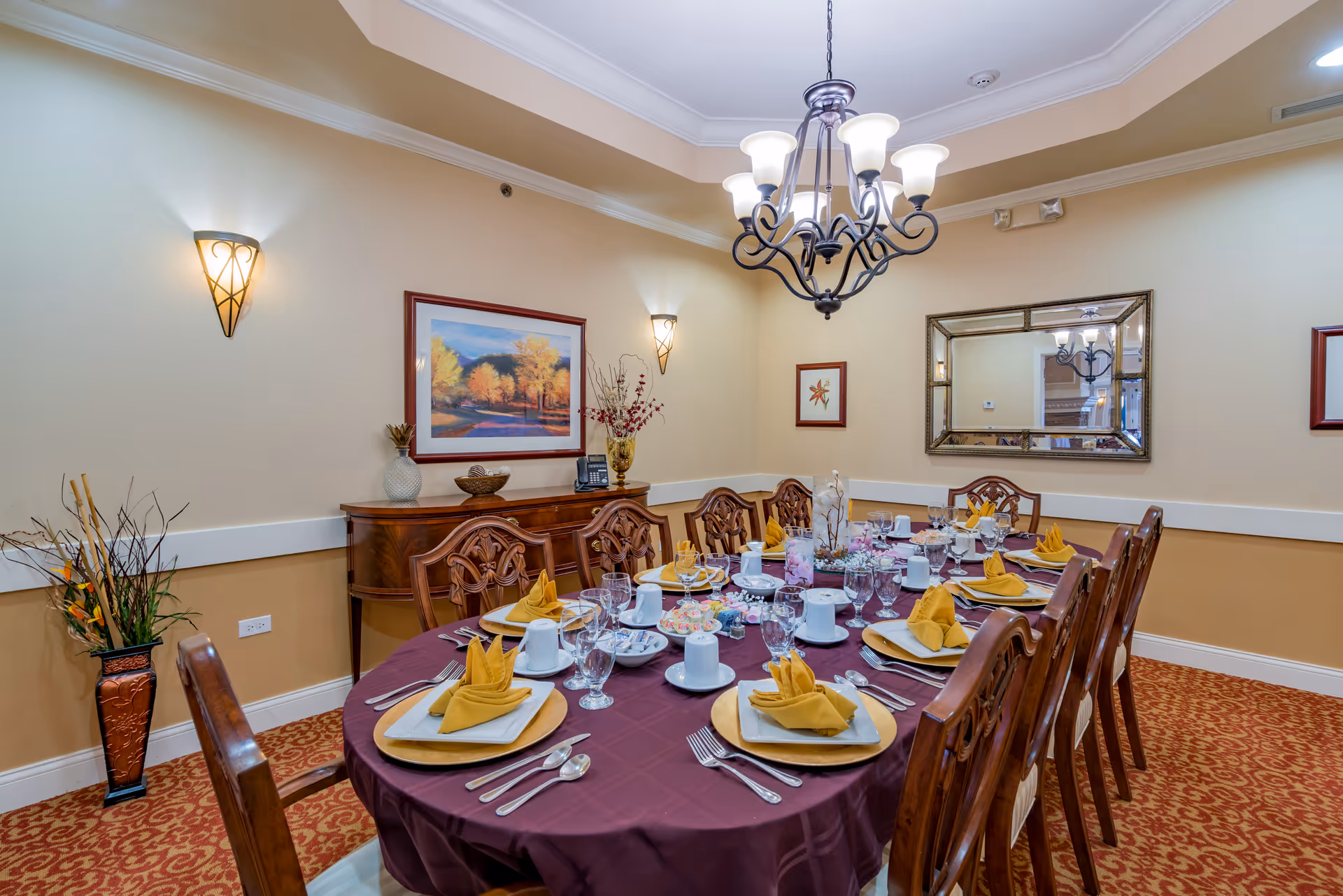 A formal dining room with a large oval table covered with a burgundy tablecloth, set with plates, glasses, silverware, and yellow folded napkins. The room has beige walls, a patterned carpet, a chandelier hanging above the table, a large mirror on one wall, framed artwork, and a wooden sideboard with decorative items.