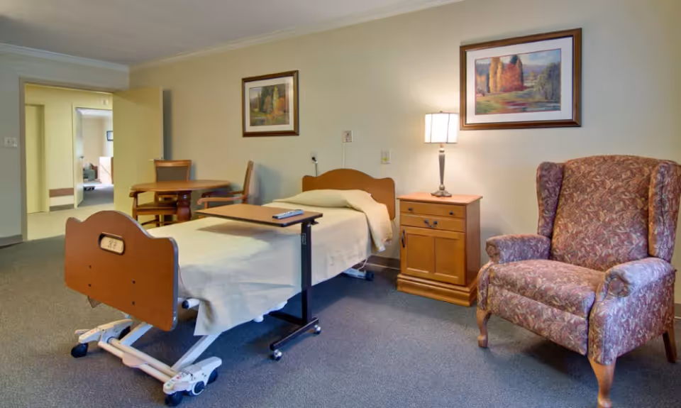 A senior living facility room with a hospital-style bed covered with a beige blanket, a wooden bedside table with a lamp, a floral upholstered armchair, and a round wooden table with chairs. Two framed landscape paintings hang on the walls.
