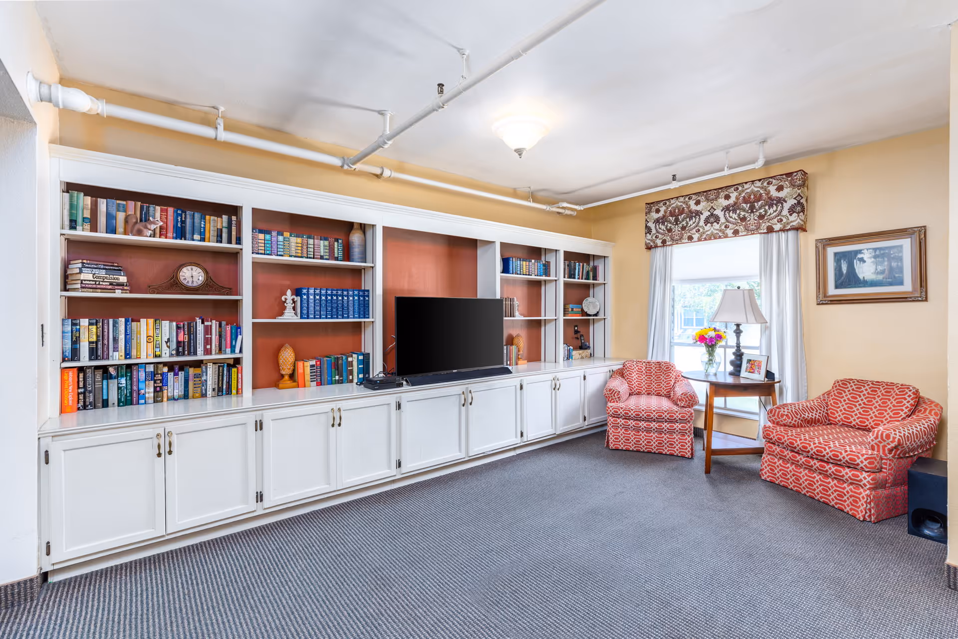 A cozy living room area with a large white built-in bookshelf filled with books and decorative items. A flat-screen TV is placed in the center of the bookshelf. Two red patterned armchairs are positioned near a window with white curtains and a floral valance. A small wooden table with a lamp, a vase of flowers, and a framed photo sits between the chairs. The walls are painted yellow, and the floor is carpeted in a gray pattern.