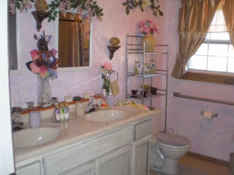 Bathroom with a double sink vanity, decorated with flowers and various toiletries. There is a toilet with a cushioned seat cover, a metal shelving unit holding decorative items, a window with brown curtains, and a toilet paper holder on the wall.