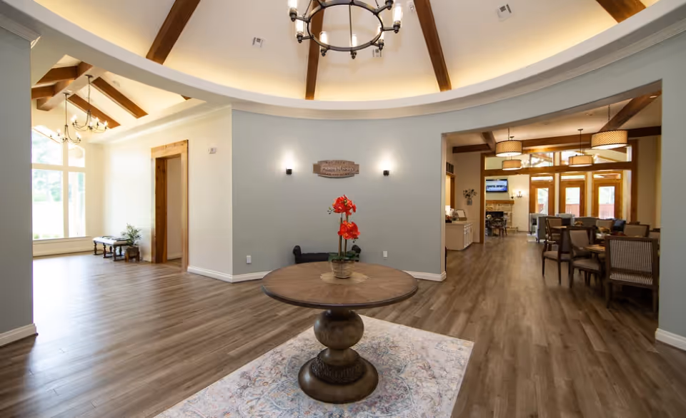 Open, bright senior living common area with a round entry table topped by red flowers, exposed wood beams, chandeliers, and dining seating visible to the right.