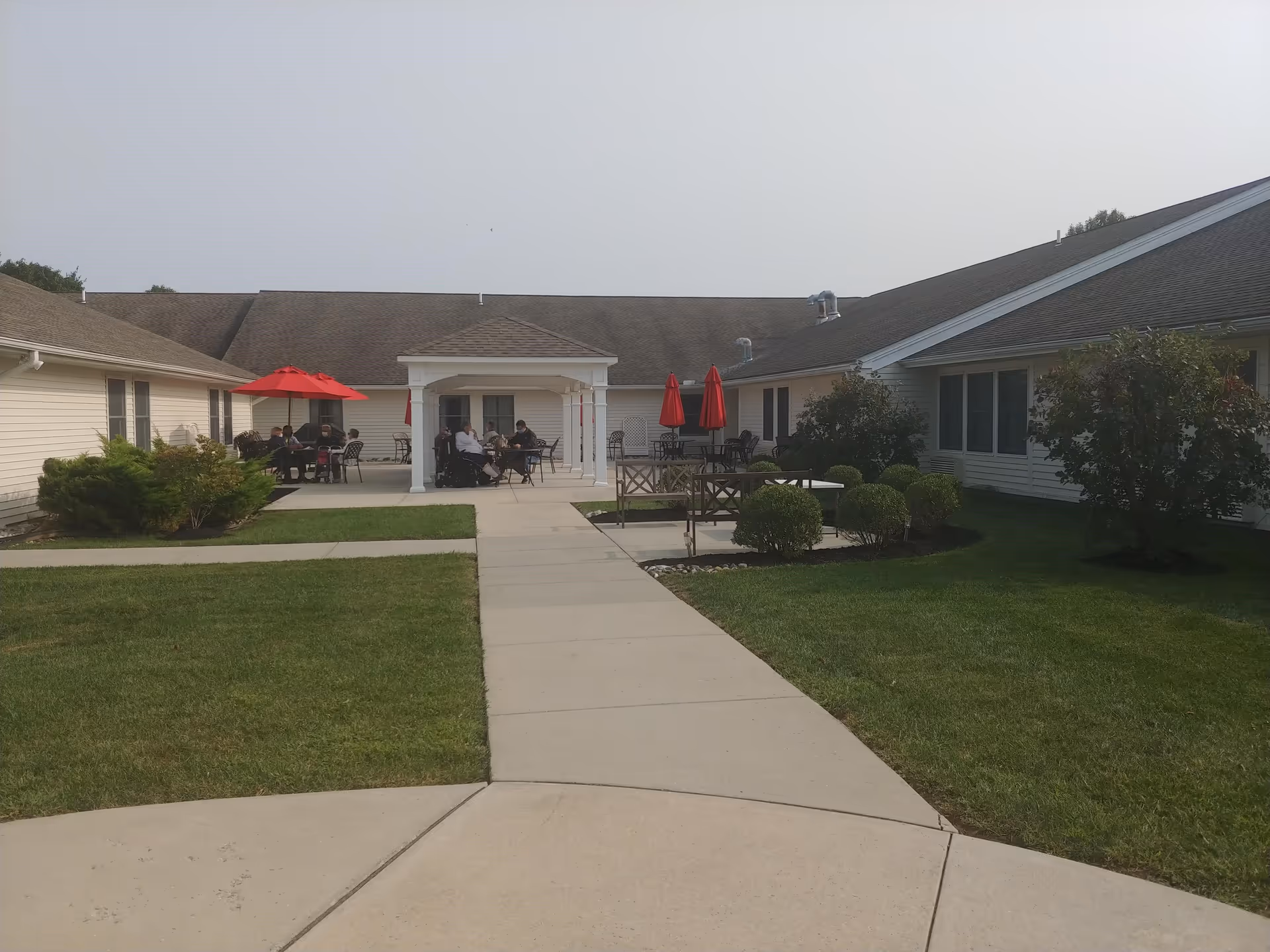 Outdoor courtyard area of a senior living facility with a concrete walkway leading to a covered seating area. Several tables with red umbrellas and chairs are arranged on the patio, with a few people sitting and socializing. The courtyard is surrounded by single-story buildings with beige siding and a well-maintained lawn with shrubs and bushes.