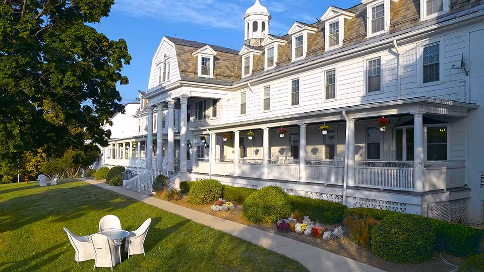 Large white multi-story building with a covered porch featuring hanging flower pots and outdoor seating. The building is surrounded by a well-maintained lawn with a small round table and four white chairs on the grass. There are bushes and flower beds along the walkway leading to the entrance. A large tree is visible on the left side under a clear blue sky.