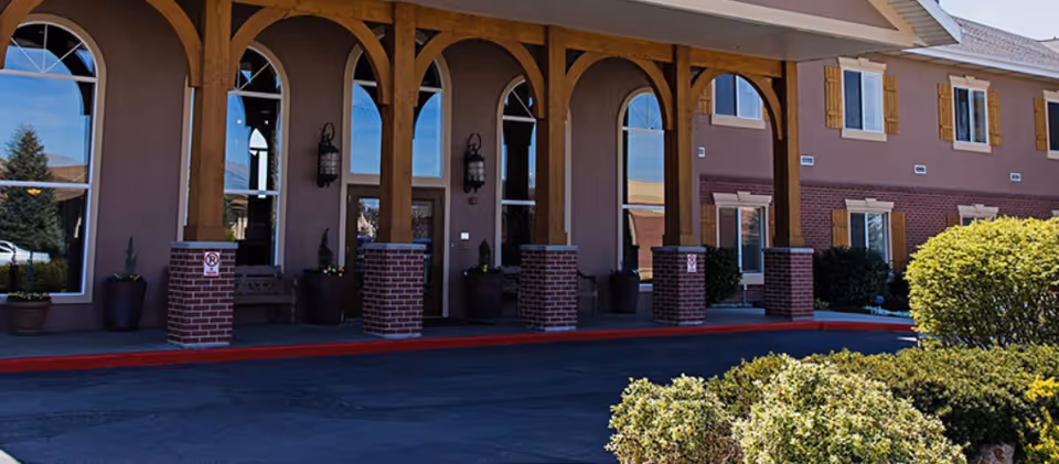 Exterior front view of Alta Ridge Assisted Living of Holladay building featuring arched windows, wooden pillars with brick bases, and a covered entrance. Shrubs and bushes are visible in the foreground along the driveway.