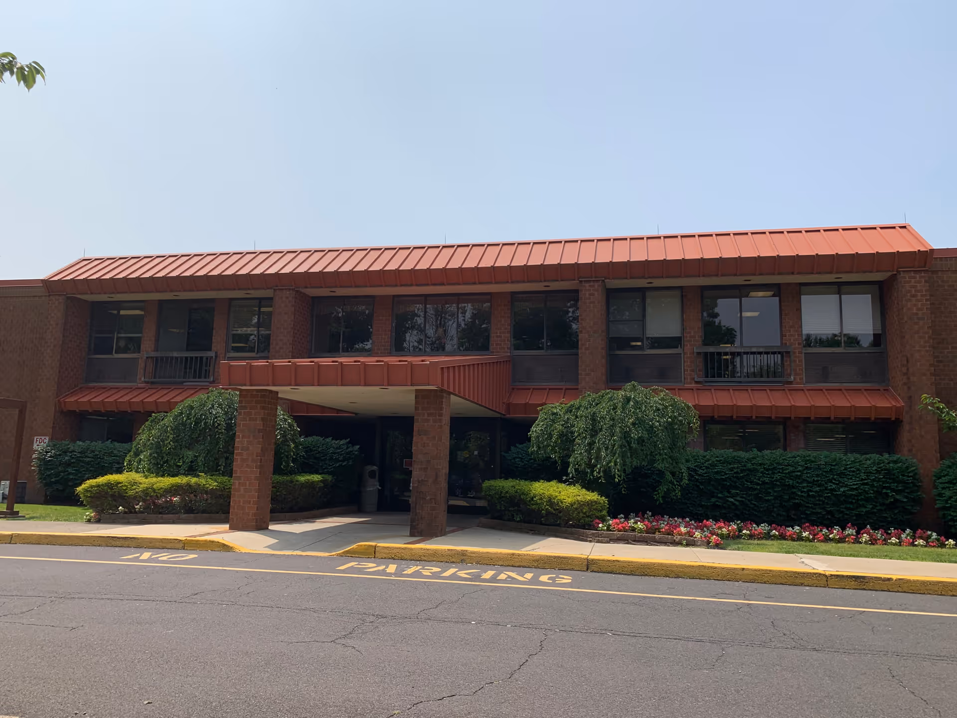 Front exterior view of River's Edge Rehabilitation and Healthcare Center, a two-story brick building with a red metal roof and entrance canopy. The entrance is flanked by neatly trimmed bushes and flower beds, with a driveway marked 'NO PARKING' in front.