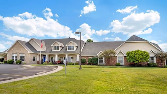 Exterior view of a single-story senior living facility building with a well-maintained lawn, bushes, and a paved driveway under a partly cloudy blue sky.
