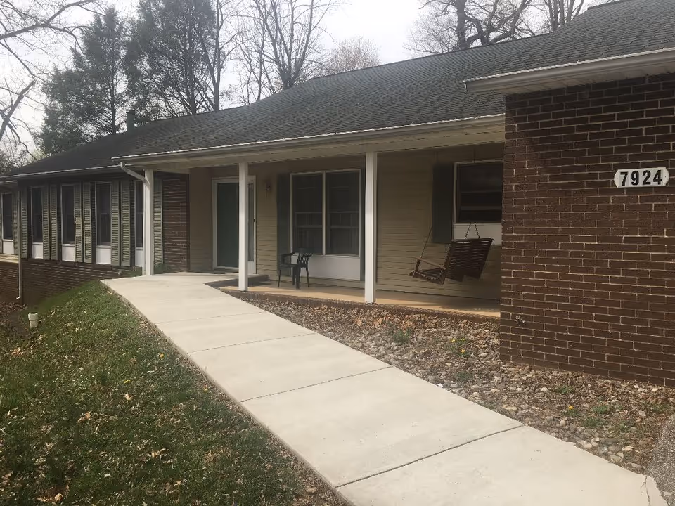 Front exterior view of a single-story brick and siding building with a covered porch, a concrete walkway leading to the entrance, a porch swing, and a plastic chair. The building number 7924 is visible on the brick wall.