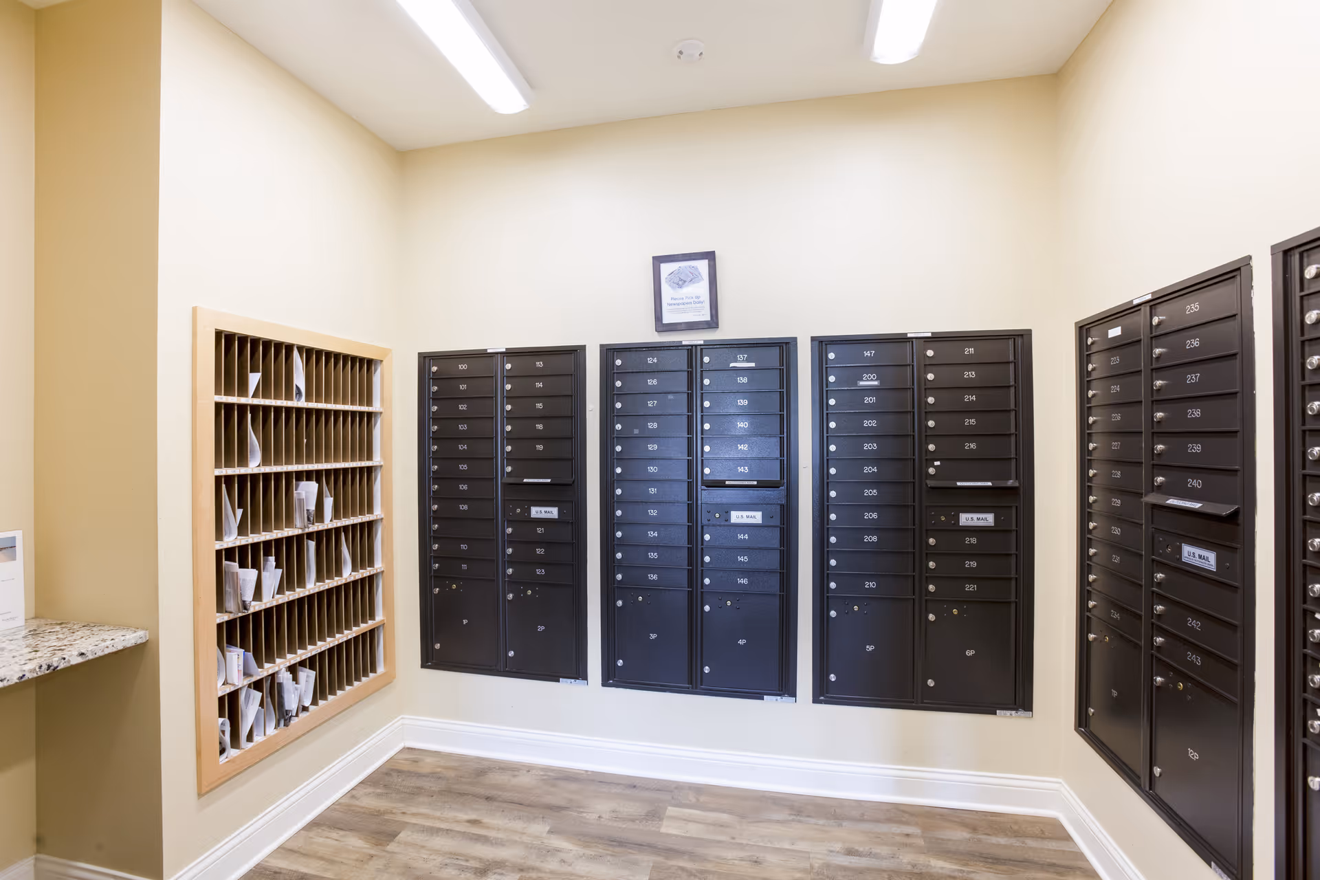Interior view of a mailroom with multiple black mailboxes mounted on beige walls and a wooden cubby shelf filled with mail. The room has wood flooring and bright overhead lighting.