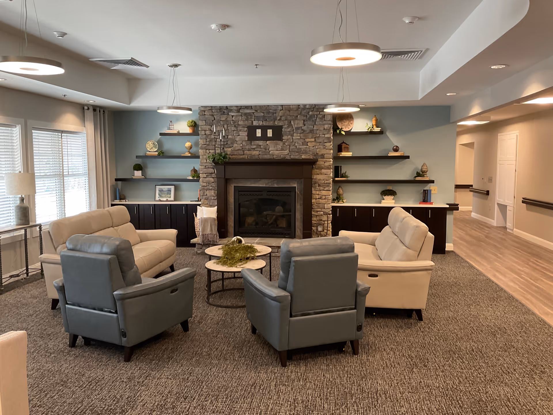 A cozy living room area in a senior living facility featuring a stone fireplace centered on the back wall with wooden shelves on either side displaying decorative items. The room has two beige sofas and two gray armchairs arranged around two round coffee tables with a plant centerpiece. Large windows with blinds and curtains allow natural light, and modern circular ceiling lights illuminate the space. The floor is carpeted with a gray pattern, and there is a hallway visible to the right with wood flooring and handrails.