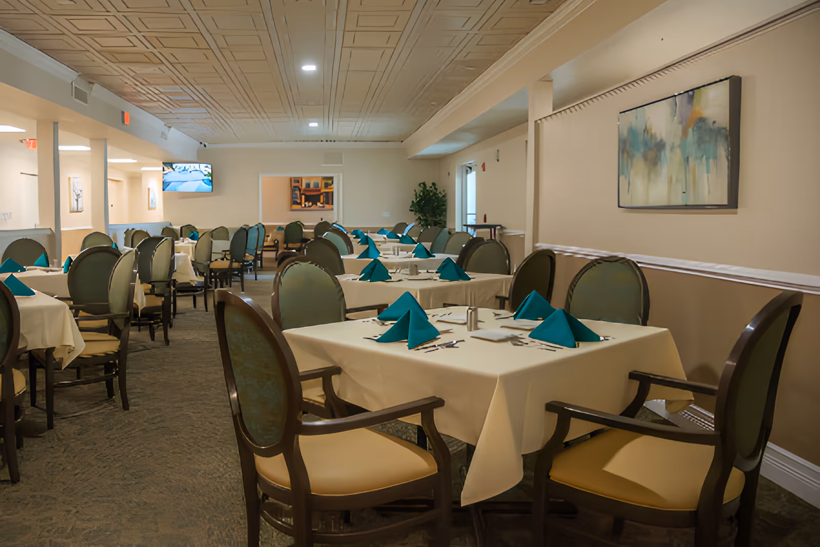 A dining room in Chateau Madeleine with multiple tables covered in white tablecloths, each set with teal folded napkins, silverware, and salt and pepper shakers. The room has green cushioned chairs with wooden frames, beige walls, a patterned ceiling, and abstract artwork on the walls. A television is mounted on the far wall.
