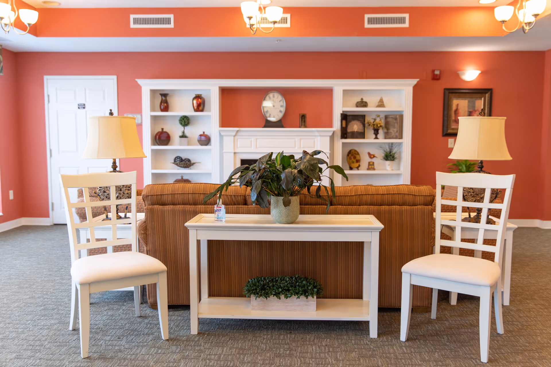 A cozy living room area with a brown striped sofa facing a white fireplace and built-in shelves decorated with vases, plants, and ornaments. In front of the sofa is a white console table with a potted plant and a small bottle. Two white chairs with cushioned seats and table lamps flank the console table. The walls are painted a warm coral color, and the carpet is gray.