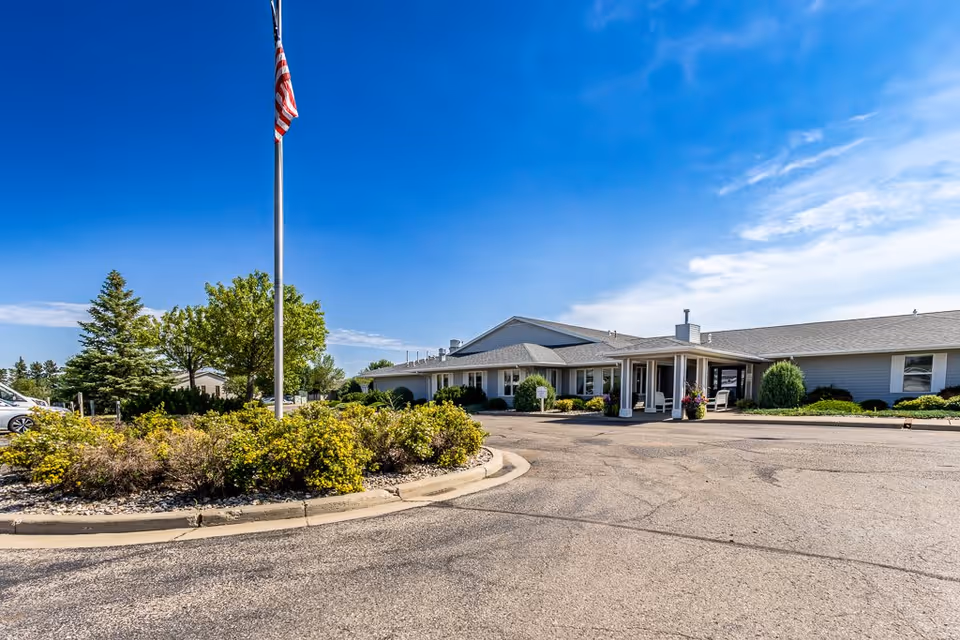 Exterior view of Elison Assisted Living of Minot facility on a clear sunny day, showing a single-story building with a covered entrance, surrounded by greenery and a flagpole with an American flag.