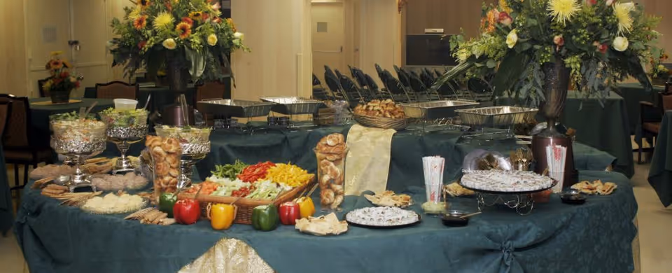 A buffet table set up with various appetizers and snacks including vegetables, crackers, and pastries, decorated with large floral arrangements in a room with chairs and tables in the background.
