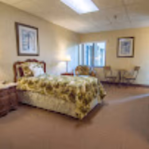 A cozy bedroom with a single bed covered in a floral bedspread, a wooden nightstand with a lamp, two chairs and a small table near a window, and framed artwork on the walls.