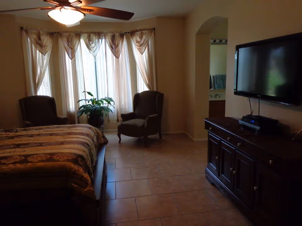 Bedroom with a bed in the foreground, two armchairs by a large curtained window, a ceiling fan, potted plant, dresser and wall-mounted TV.