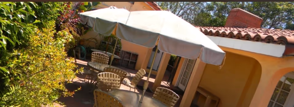 Outdoor patio area with a large white umbrella shading a glass table surrounded by woven chairs. The patio is adjacent to a building with peach-colored walls and a tiled roof, with greenery visible on the left side.