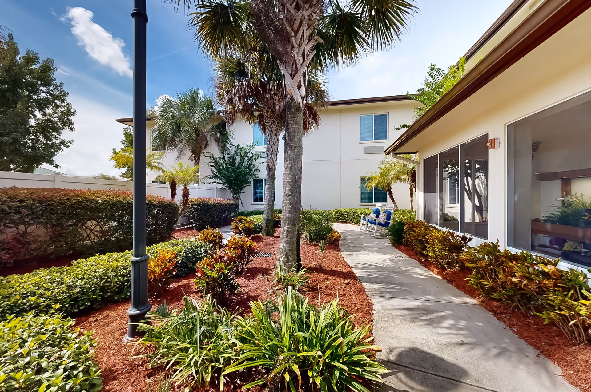 Outdoor garden area at The Windsor of Palm Coast featuring a paved walkway, palm trees, various shrubs, and two blue cushioned chairs near a building with large windows under a clear blue sky.