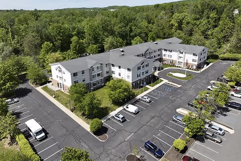 Aerial view of a three-story senior living facility named Brookdale Wayne surrounded by trees and greenery, with a large parking lot featuring multiple parked cars and vans.