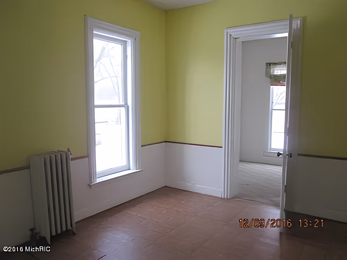 Empty room with yellow upper walls and white lower walls separated by a brown trim, a window with white trim, a white radiator, and an open door leading to another room with a window.