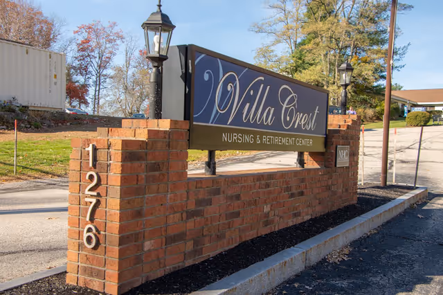 A brick sign at the entrance of Villa Crest Nursing and Retirement Center with the address number 1276 displayed vertically on the left side. The sign is flanked by two black lamp posts and there are trees and a building visible in the background under a clear blue sky.