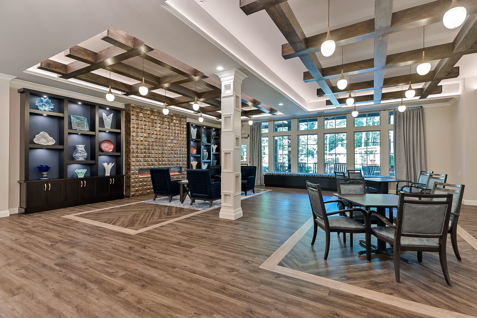 Spacious and modern senior living common area with wood flooring, decorative ceiling beams, pendant lights, a seating area with armchairs around a fireplace, display shelves with decorative glass pieces, and tables with chairs near large windows letting in natural light.