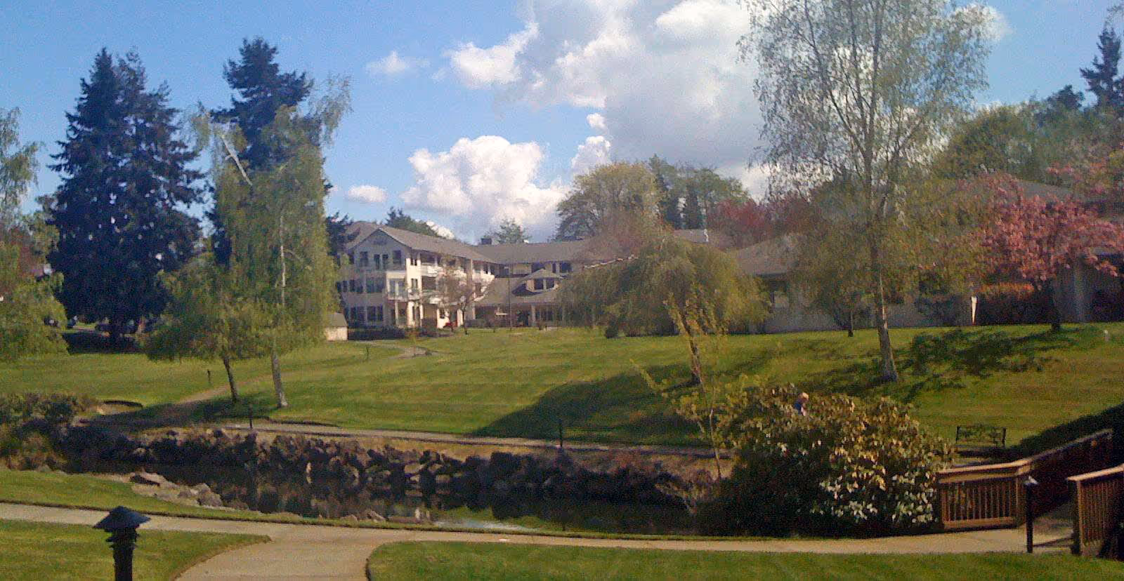 View of a senior living facility building surrounded by a large green lawn with trees and bushes under a partly cloudy sky. A paved walkway and wooden railings are visible in the foreground.