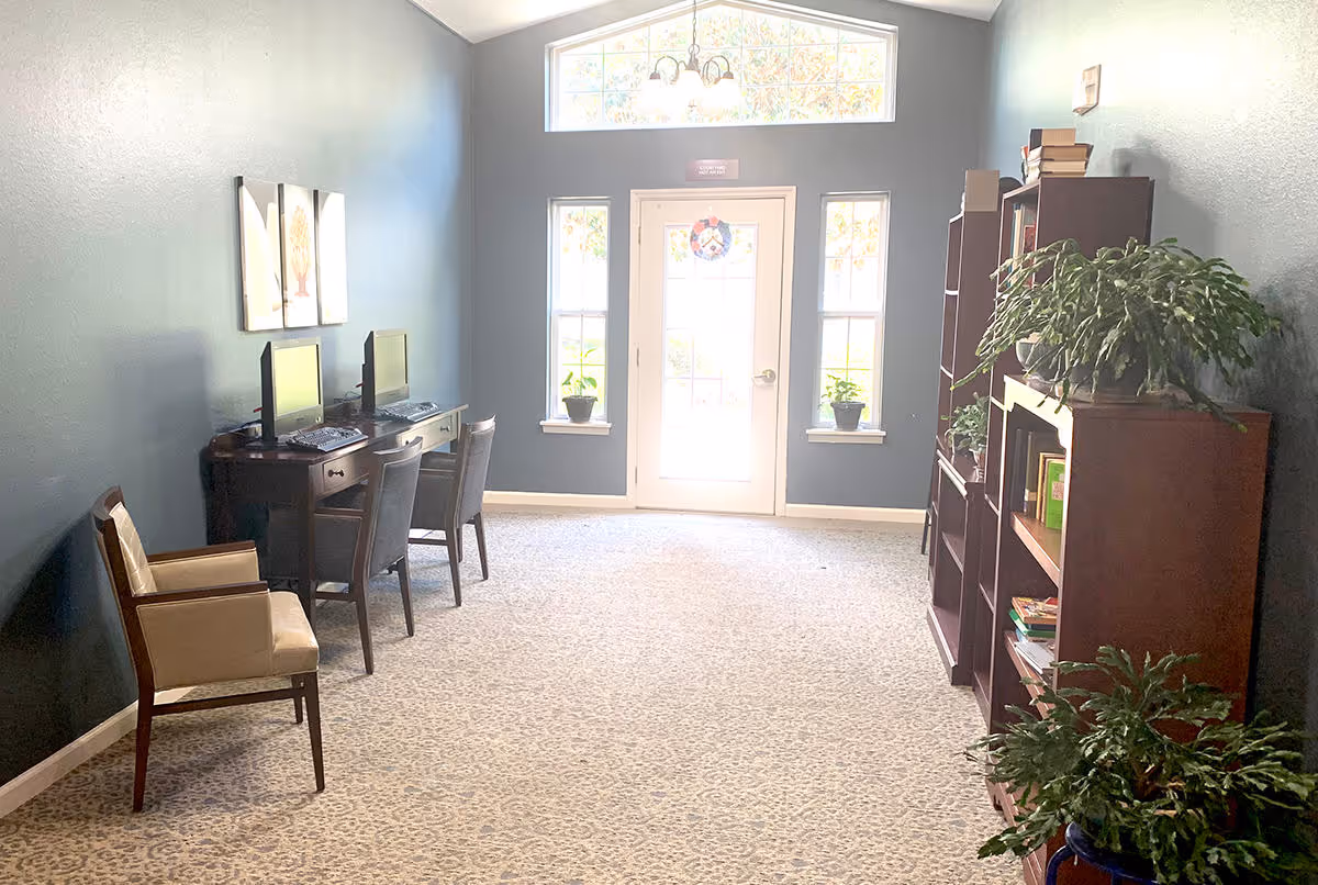 A bright room with blue walls and carpeted floor featuring two computer desks with chairs on the left side, a single armchair, and two tall bookshelves with plants and books on the right side. A door with windows on either side and a transom window above lets in natural light at the far end of the room.