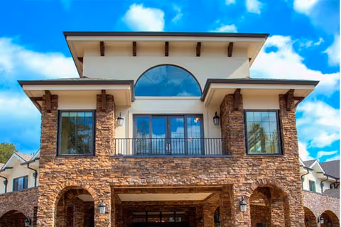 Front façade of a stone-clad multi-story assisted living building with arched entrance, balcony, and large windows under a blue sky.