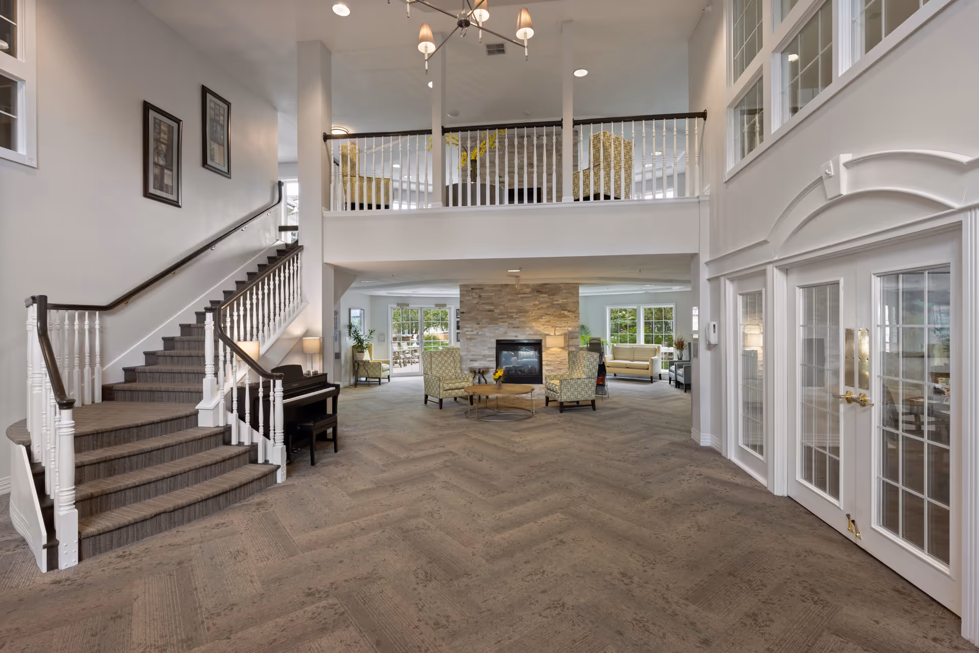 Spacious and well-lit interior common area of a senior living facility with a staircase on the left, a piano beneath it, a stone fireplace in the center surrounded by comfortable chairs and sofas, large windows letting in natural light, and a second-floor balcony with additional seating visible above.