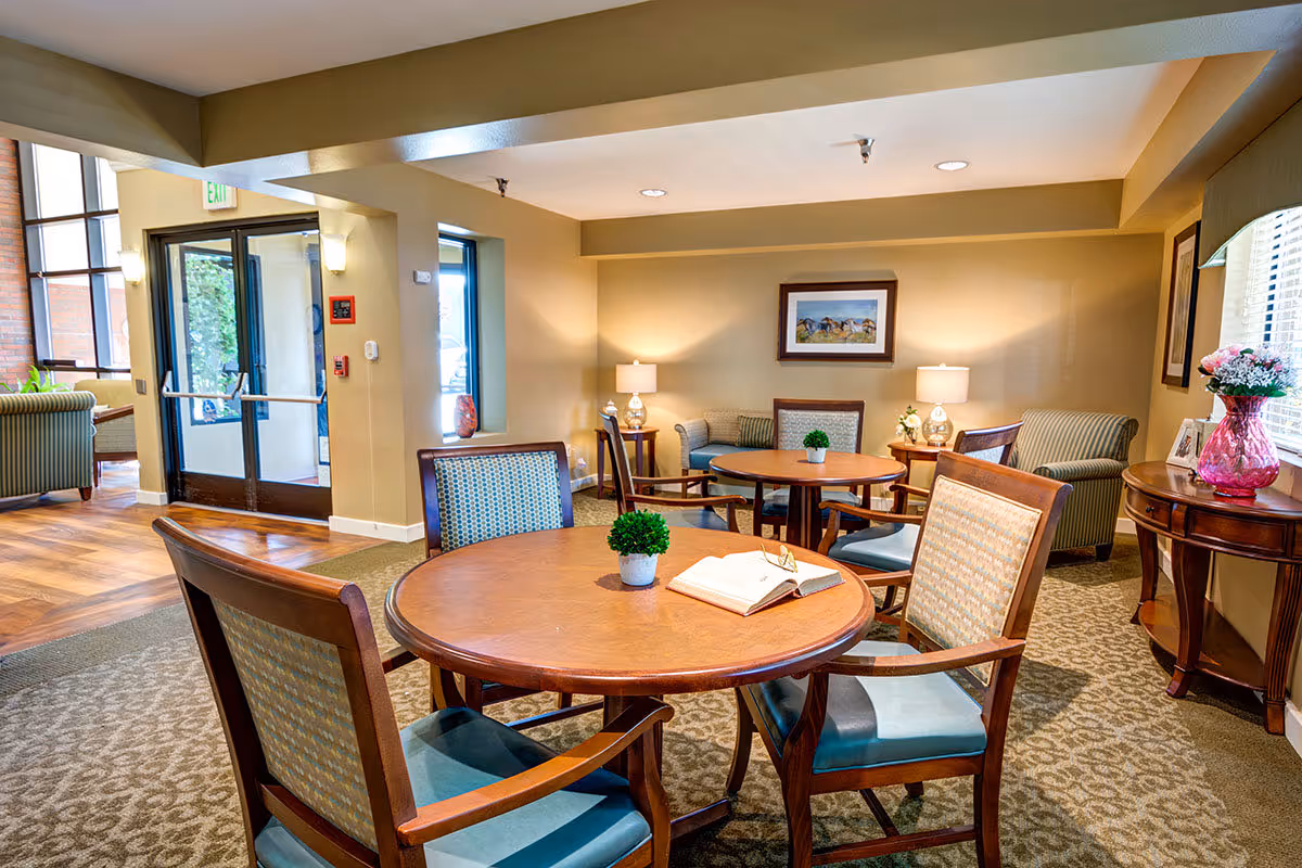 A cozy common area in a senior living facility featuring round wooden tables with chairs, a small potted plant and an open book on the nearest table. The room has beige walls, carpeted floors, two table lamps on side tables, framed artwork on the walls, and a window with blinds and a vase of flowers on a wooden console table. In the background, there is a glass door entrance and additional seating with armchairs.
