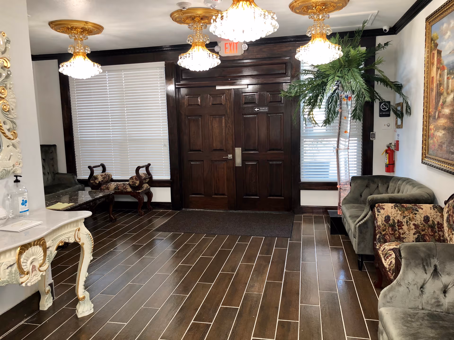 An elegant interior lobby area with dark wooden double doors, three ornate chandeliers hanging from the ceiling, a green velvet sofa, floral upholstered chairs, a large potted artificial palm tree, a decorative white and gold console table with hand sanitizer on it, and a large framed painting on the wall.