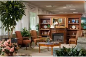 Communal lounge with armchairs, a coffee table, indoor plants and a decorative fireplace shelving unit in front of large windows.
