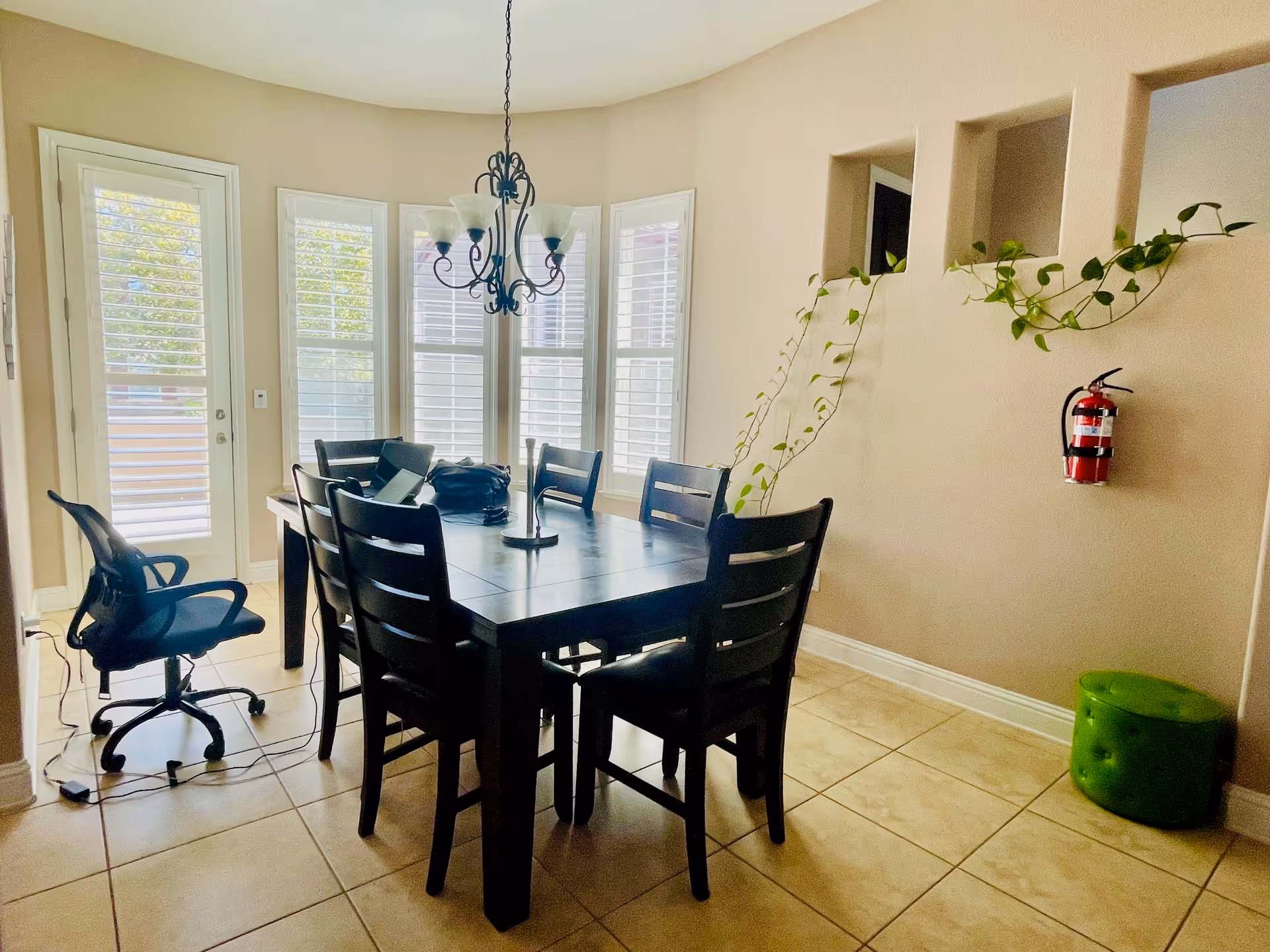 A dining area with a dark wooden table surrounded by six chairs, including one office chair on wheels. The room has large windows with white shutters allowing natural light to enter. A chandelier with five lights hangs above the table. There is a fire extinguisher mounted on the beige wall, a green ottoman on the floor, and a trailing plant growing through three rectangular wall openings.