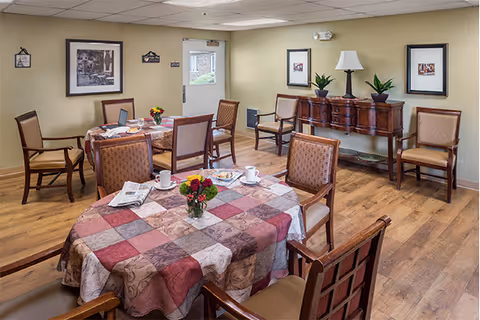A dining room in a senior living facility with tables covered in patterned tablecloths, each set with cups, plates, and small flower arrangements. The room has wooden chairs, framed pictures on the walls, a wooden sideboard with plants and a lamp, and a wood floor.