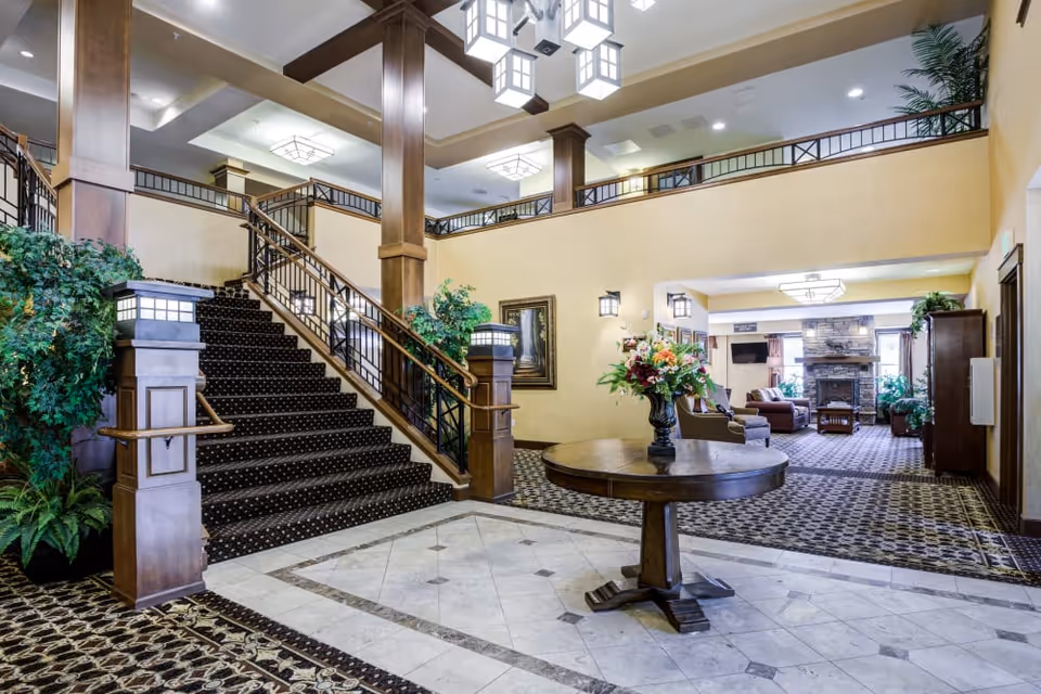 Spacious and elegant interior of The Lodge at Mallard's Landing featuring a grand staircase with dark carpet and wooden railings, a round wooden table with a floral arrangement in the center, patterned carpet flooring, and a cozy seating area with a stone fireplace and TV in the background. The space is well-lit with modern ceiling lights and decorated with plants and framed artwork.