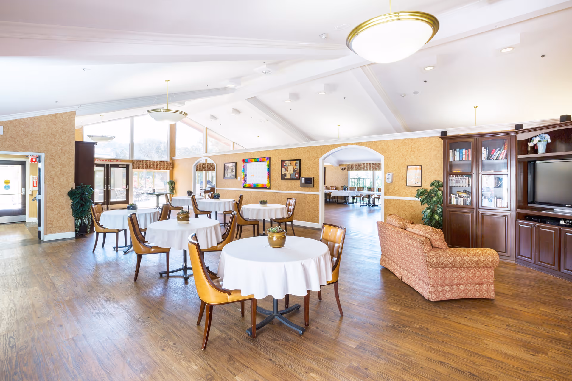 A bright and spacious common area in a senior living facility with round tables covered in white tablecloths, surrounded by wooden chairs with leather seats. There is a patterned sofa facing a large wooden entertainment center with a TV and bookshelves. The room has high vaulted ceilings with modern light fixtures, large windows letting in natural light, and wooden flooring. The walls are decorated with framed pictures and a colorful bulletin board. An adjacent room with more tables is visible through an arched doorway.