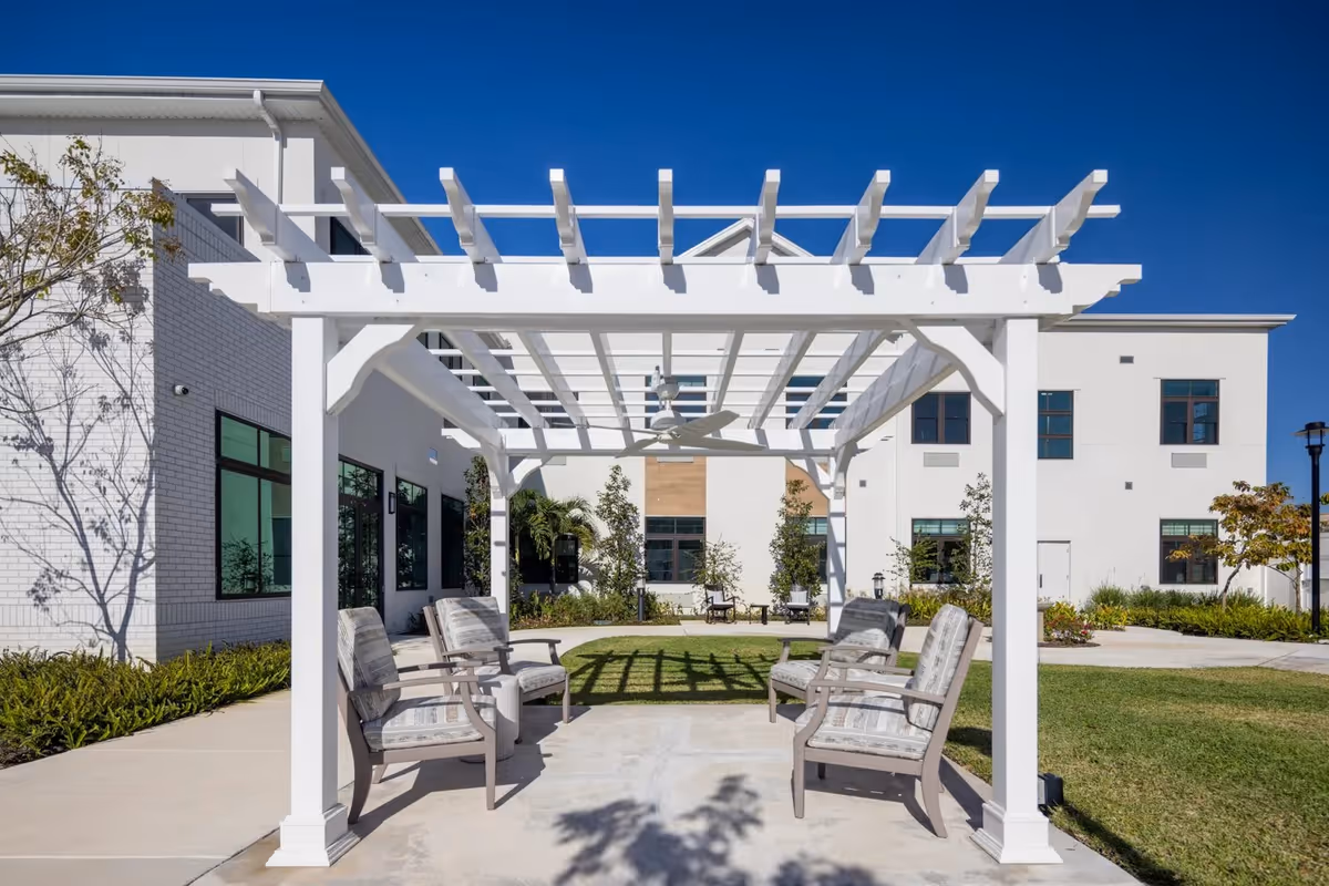 Outdoor seating area at American House Boynton Beach featuring a white pergola with four cushioned chairs arranged underneath on a concrete patio, surrounded by green grass, trees, and a white building in the background under a clear blue sky.