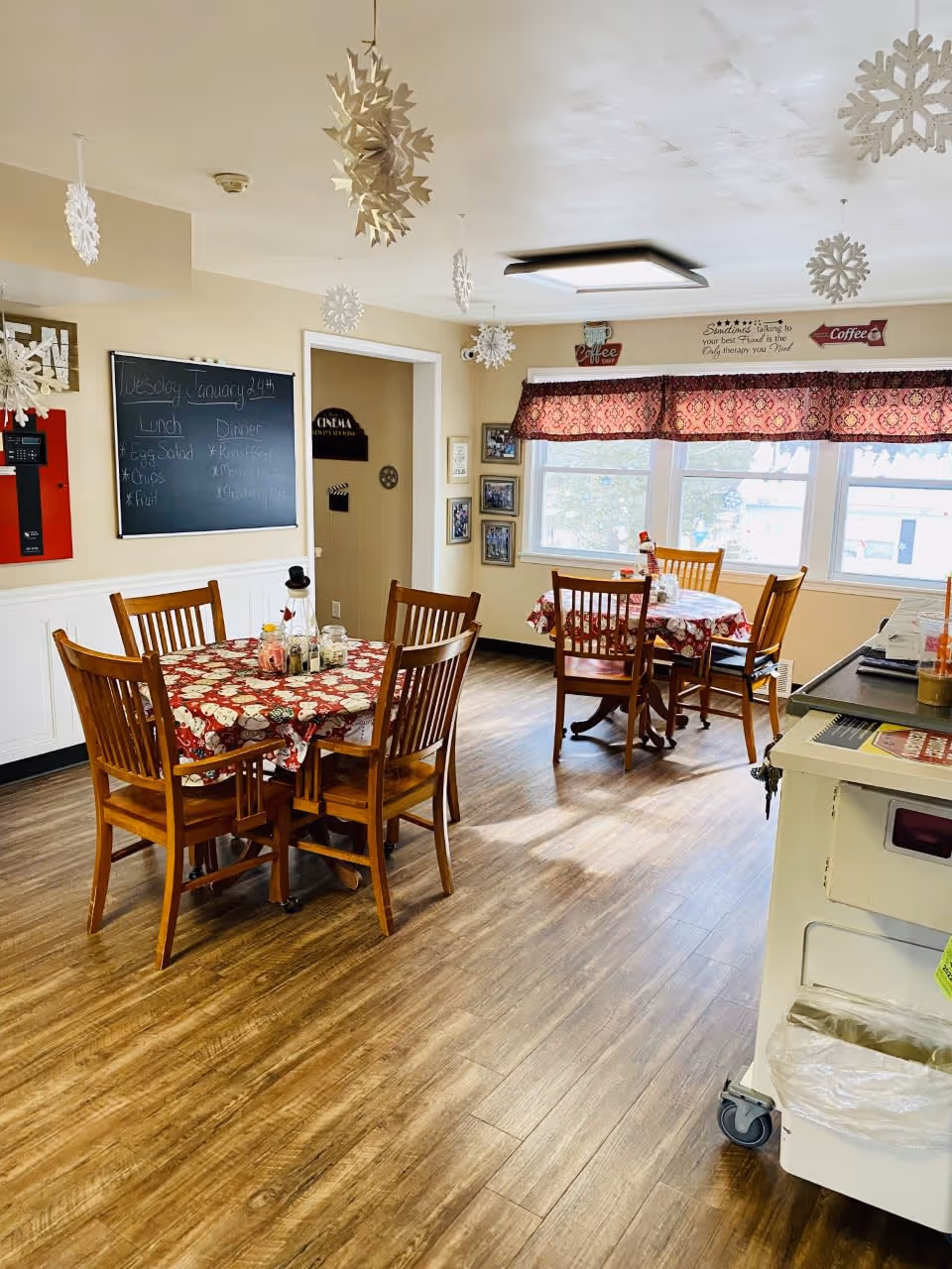 A cozy dining room with wooden floors and two round tables covered with red and white patterned tablecloths. Each table is surrounded by wooden chairs. Snowflake decorations hang from the ceiling. A chalkboard on the wall displays the lunch and dinner menu for Tuesday, January 24th. A window with a red patterned valance lets in natural light. Various wall decorations include coffee signs and framed pictures.