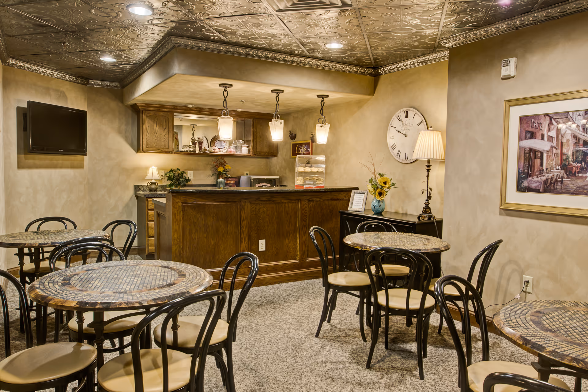 A cozy dining area with several round mosaic-tiled tables and black chairs with beige cushions. In the background, there is a wooden counter with three hanging pendant lights above it, a small lamp, and decorative items including flowers and a clock on the wall. A flat-screen TV is mounted on the left wall, and a framed painting hangs on the right wall.