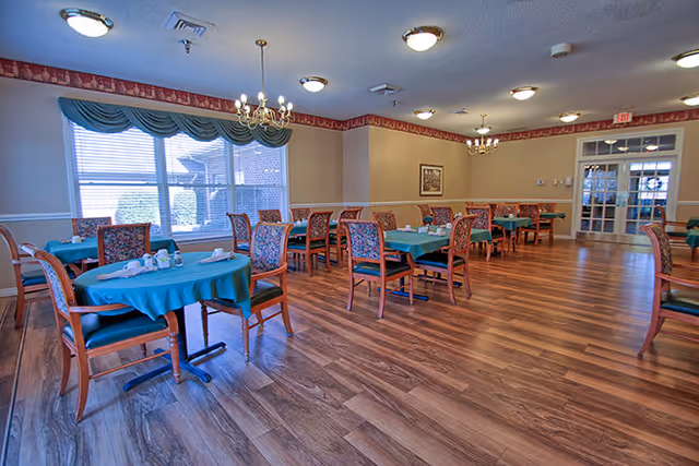 A dining room with several tables covered in teal tablecloths and surrounded by wooden chairs with patterned upholstery. The room has wood flooring, large windows with green valances, chandeliers, and soft lighting. There is a door with glass panels at the far end of the room.