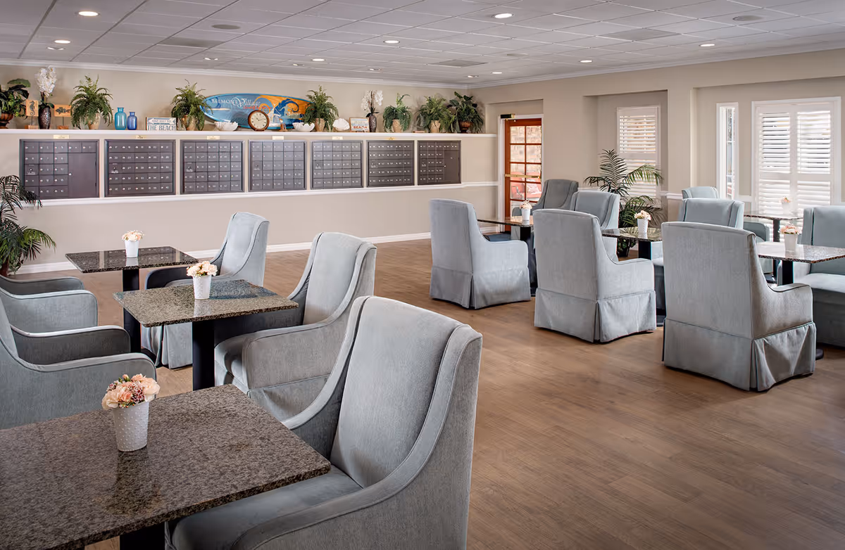 A bright and spacious common area with multiple tables and comfortable upholstered chairs arranged neatly. The room features a wooden floor, large windows with white shutters, and a wall of mailboxes decorated with plants and beach-themed decor above.