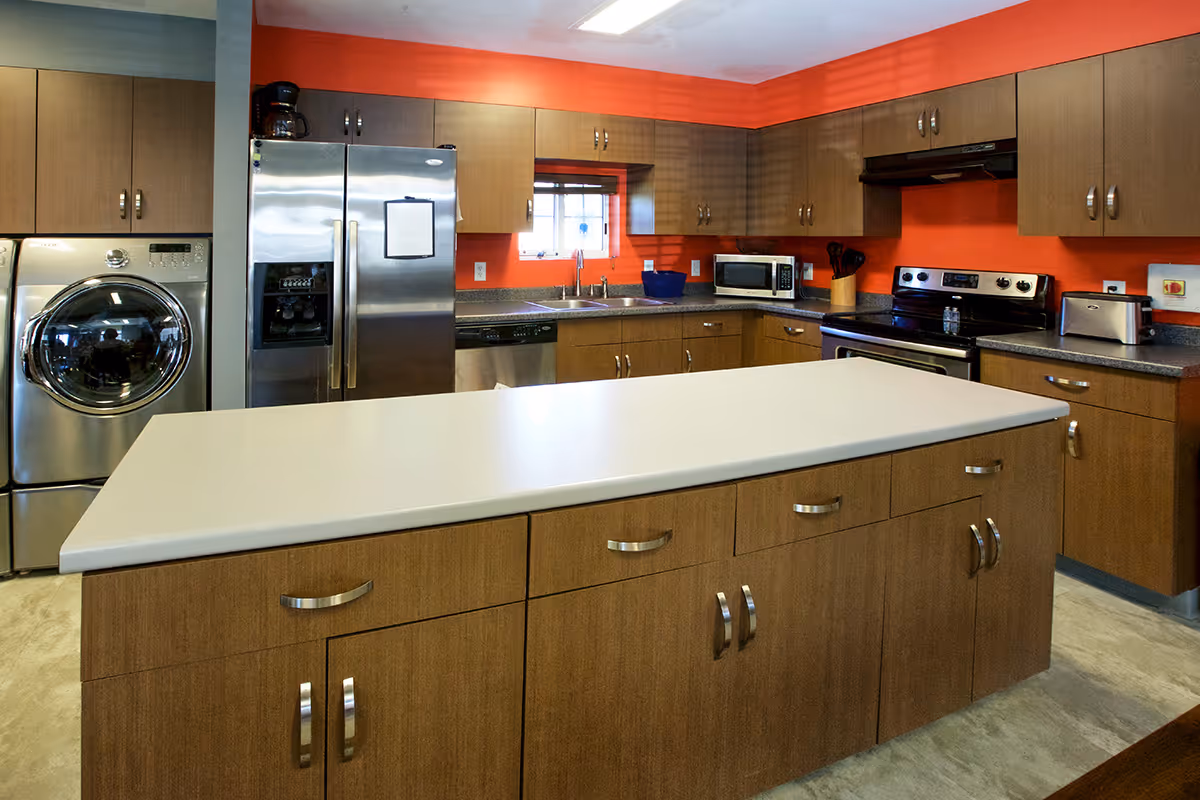 Modern kitchen with wooden cabinets, a large white island countertop in the center, stainless steel refrigerator, stove, microwave, toaster, dishwasher, and a washing machine. The walls are painted orange with a small window above the sink.