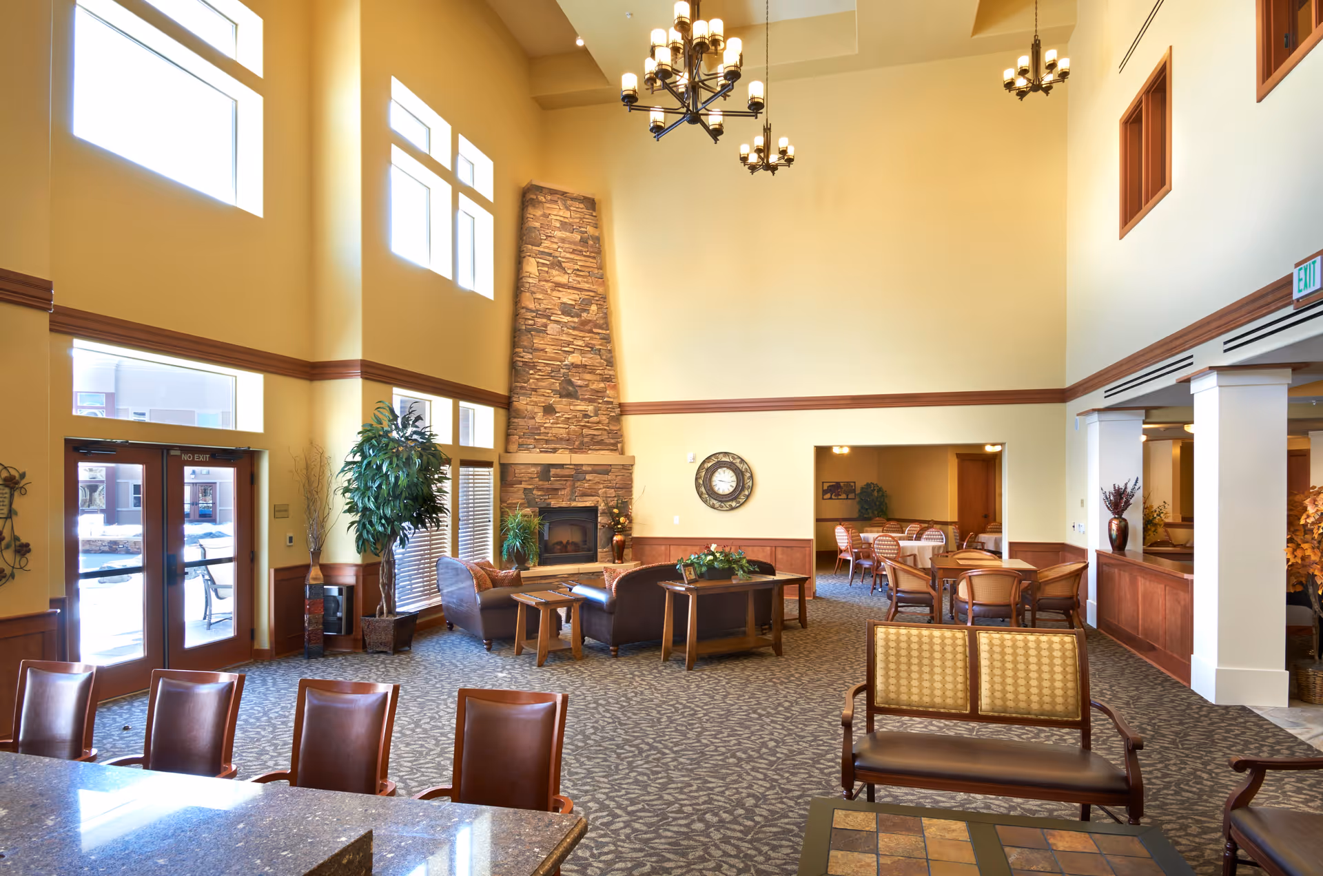 Spacious senior living facility common area with high ceilings, large windows, a stone fireplace, multiple seating arrangements including leather sofas, wooden chairs, and tables, and decorative plants. The room is well-lit with chandeliers and natural light from the windows.
